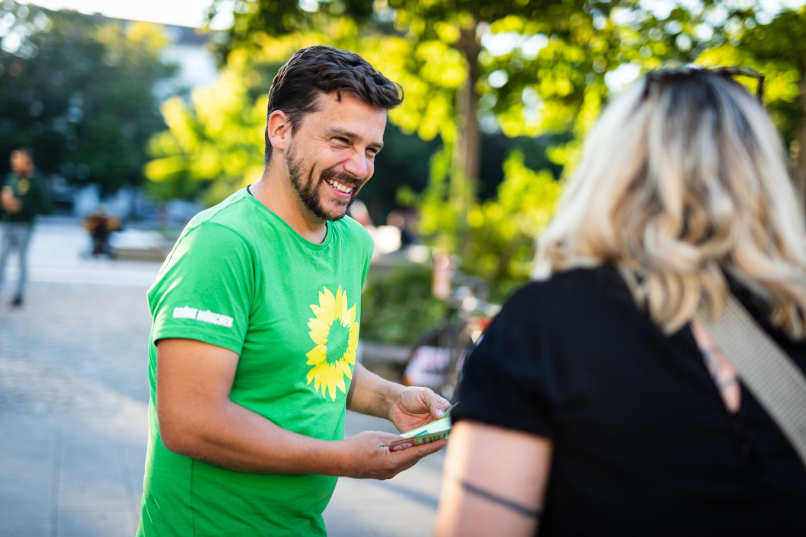 Am grünen Infostand: Eines unserer Mitglieder, ein junger Mann im grünen T-Shirt. überreicht gerade lachend einen Flyer an eine Passantin