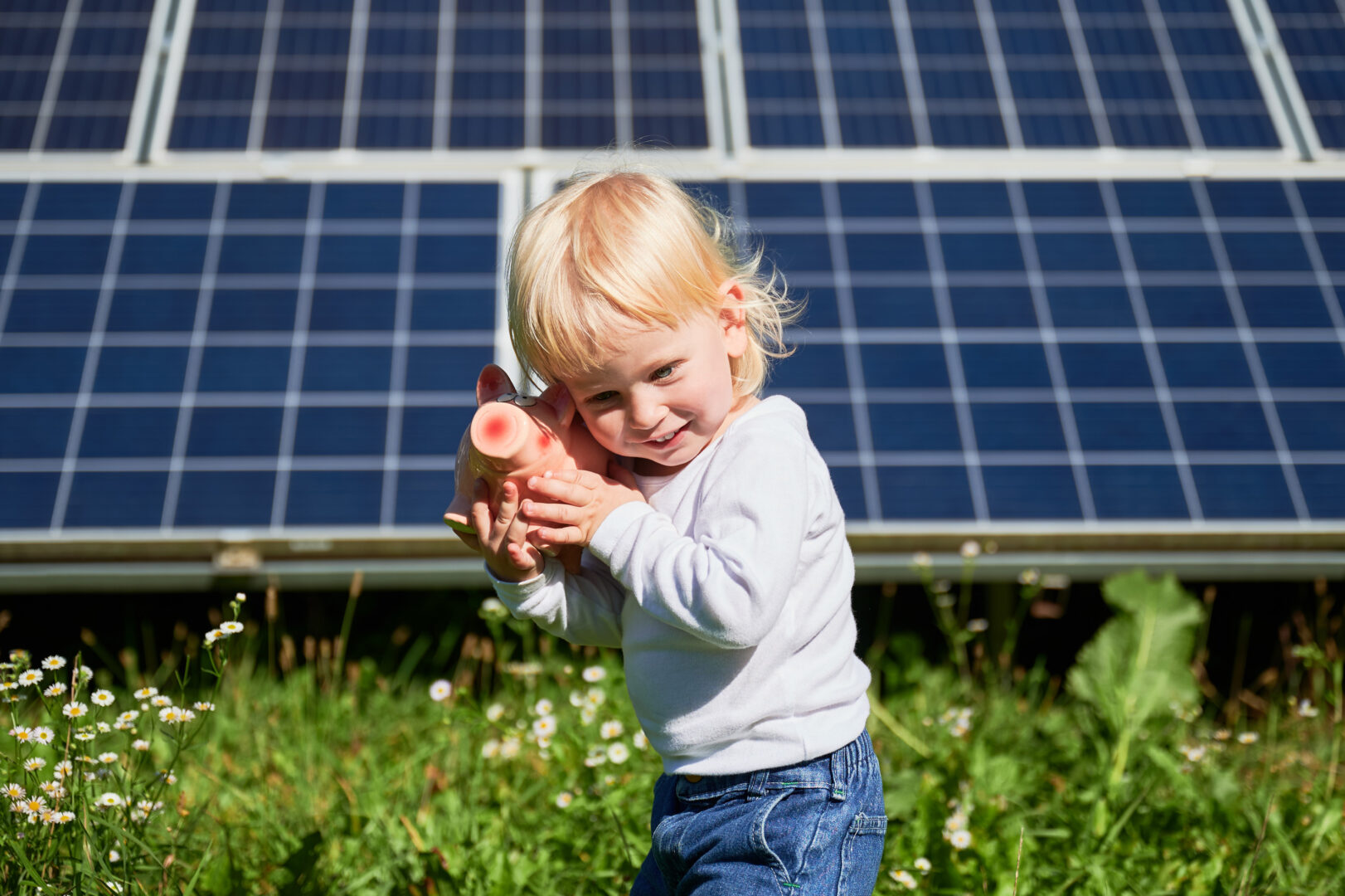 Ein kleines Kind steht vor einer Solaranlage inmitten einer Blumenwiese. Es hält lächelnd ein Sparschwein ans Ohr. Offenbar schüttelt es das Sparschwein gerade, um abzuschätzen, wie viel Geld wohl darin ist.